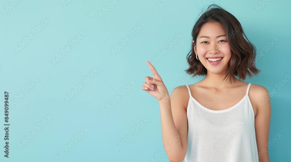 An attractive Asian model pointing with a smile, standing against a serene light blue background with ample copy space. An attractive Asian model pointing with a smile, standing against a serene light blue background with ample copy space.