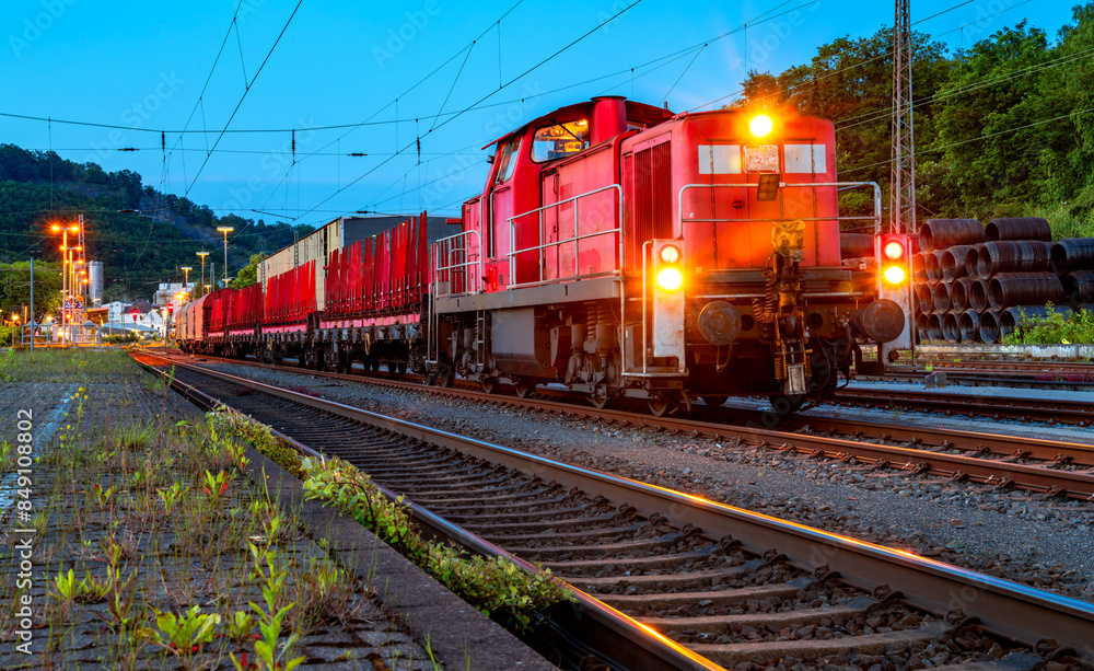 Obraz premium Red freight train with old powerful diesel engine waiting for departure at Letmathe station in Germany. Evening twilight atmosphere with bright lights at blue hour. Locomotive with headlights on.