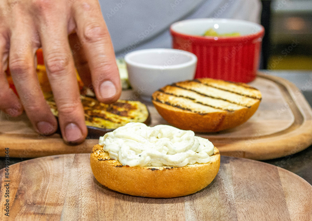 Chef hands starting the preparation of a vegetarian hamburger with bread, eggplant and sausage. Burger prepared on a restaurant kitchen. Professional chef is preparing burger. Burger making. Chef hands starting the preparation of a vegetarian hamburger with bread, eggplant and sausage. Burger prepared on a restaurant kitchen. Professional chef is preparing burger. Burger making.