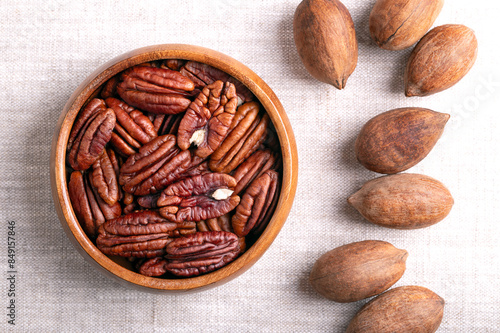 Pecan nuts in a wooden bowl on linen fabric. Dried pecan halves and whole pecans in shells, seeds and nuts of Carya illinoinensis. Ready to eat as snack, and also used for baking. Close-up from above. photo