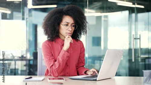 Thoughtful concentrated young african american female employee typing on laptop and thinking about problem solving at workplace in office. Serious black woman is working in computer application