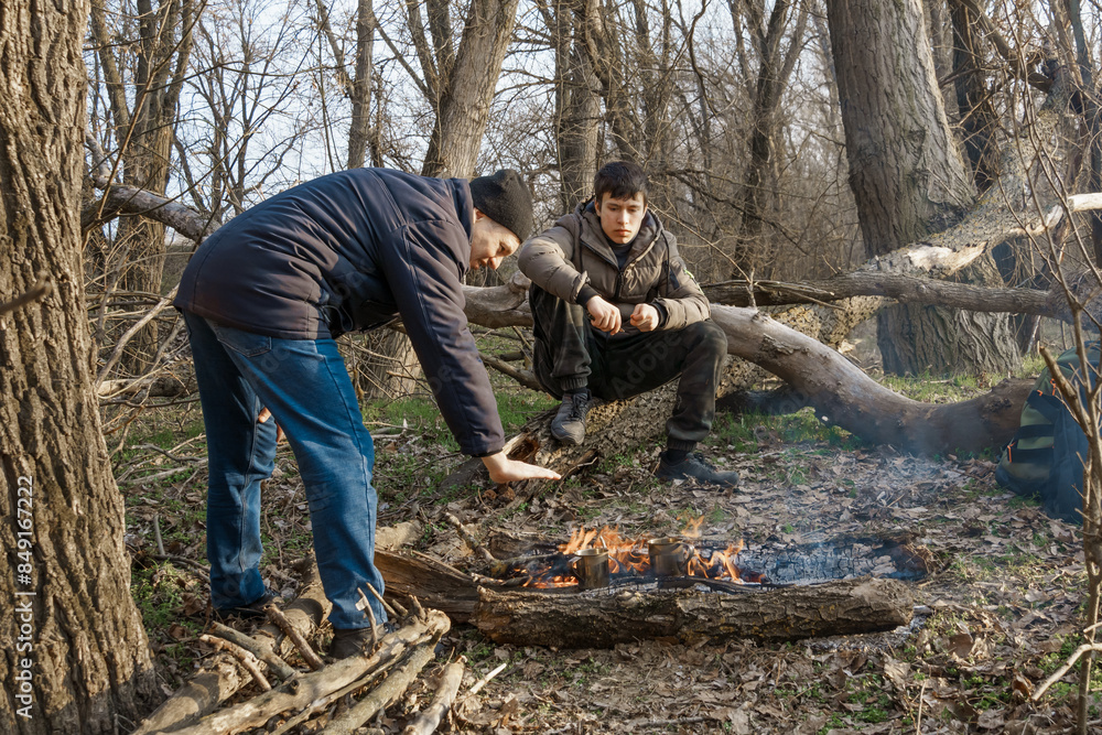 Two men make a campfire for cooking in the forest, father and son sitting on a log while hiking and outdoor activities, early spring landscape Two men make a campfire for cooking in the forest, father and son sitting on a log while hiking and outdoor activities, early spring landscape