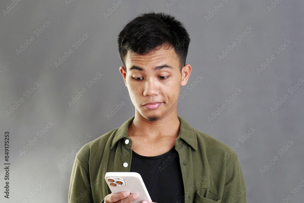 Portrait of young Asian man holding smartphone and looking at it with cynical and suspicious gaze with raised eyebrows and lips Portrait of young Asian man holding smartphone and looking at it with cynical and suspicious gaze with raised eyebrows and lips