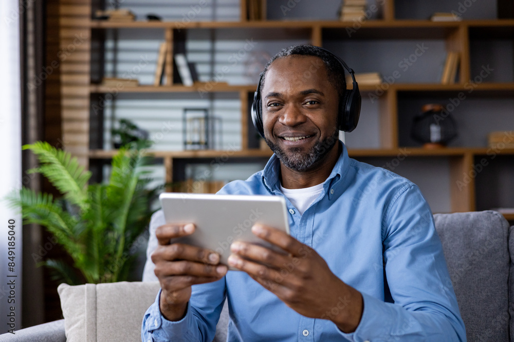 Happy man wearing headphones using a tablet while sitting in a cozy living room environment. The setting includes bookshelves and greenery, suggesting a relaxed and comfortable atmosphere. Happy man wearing headphones using a tablet while sitting in a cozy living room environment. The setting includes bookshelves and greenery, suggesting a relaxed and comfortable atmosphere.