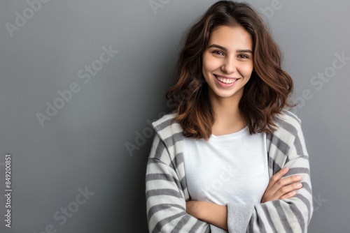 Portrait of a young latin woman with pleasant smile and crossed arms isolated on grey wall with copy space. Beautiful girl with folded arms looking at camera against grey wall. Cheerful hispanic woman