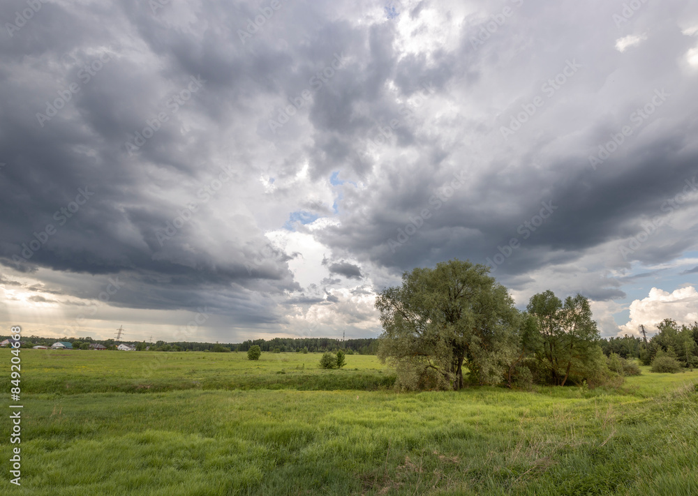 A picturesque view of a lush green field with scattered trees, set against a dramatic spring sky filled with dark, brooding clouds. A picturesque view of a lush green field with scattered trees, set against a dramatic spring sky filled with dark, brooding clouds.