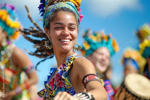 A smiling woman dances with vibrant energy at a colorful community festival. The woman wears a colorful head wrap and traditional clothing, surrounded by other dancers
