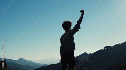 Silhouette of a person with raised fist standing on a hilltop, embracing the scenic view of mountains against a clear blue sky. photo