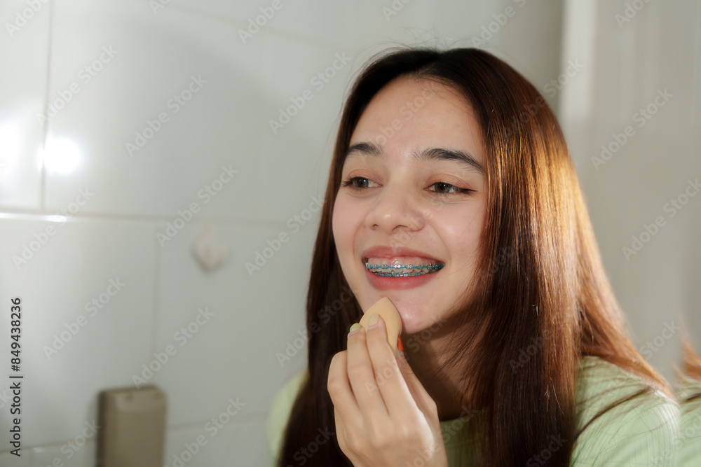 Asian woman with braces applying foundation with sponge in front of mirror. Indoor setting, natural light. Smiling, casual moment emphasizing beauty routine and self-care. Asian woman with braces applying foundation with sponge in front of mirror. Indoor setting, natural light. Smiling, casual moment emphasizing beauty routine and self-care.