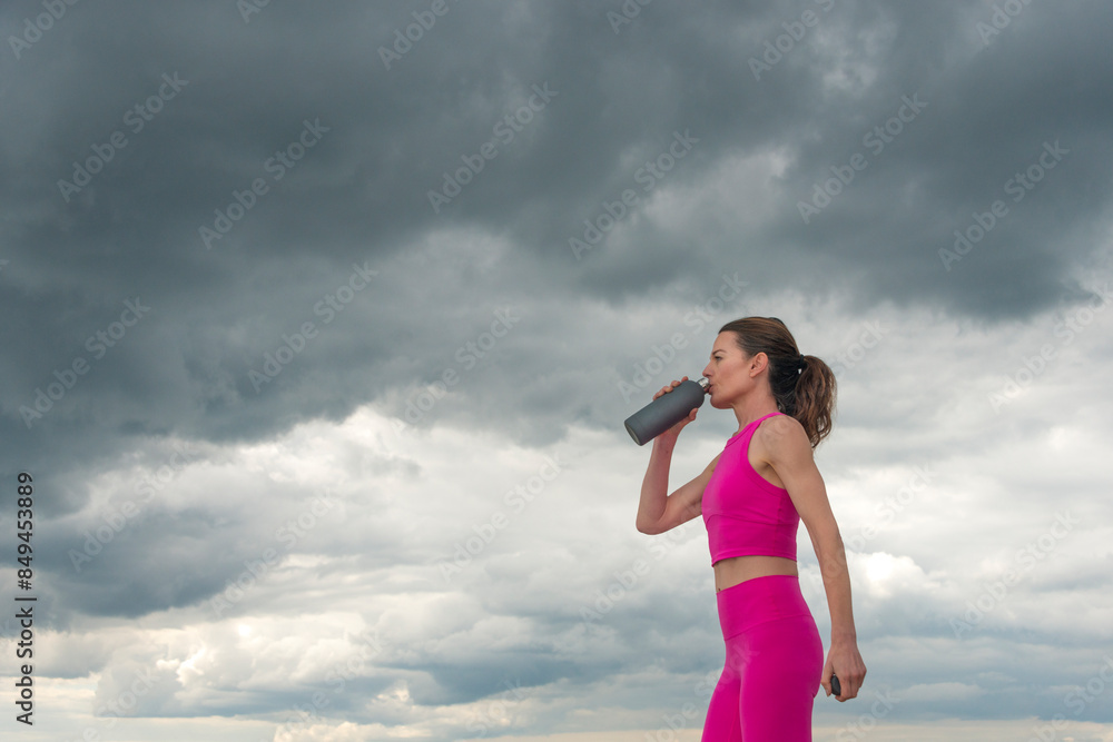 Sporty woman drinking water from a bottle, wearing pink sports clothing. dramatic sky. Sporty woman drinking water from a bottle, wearing pink sports clothing. dramatic sky.