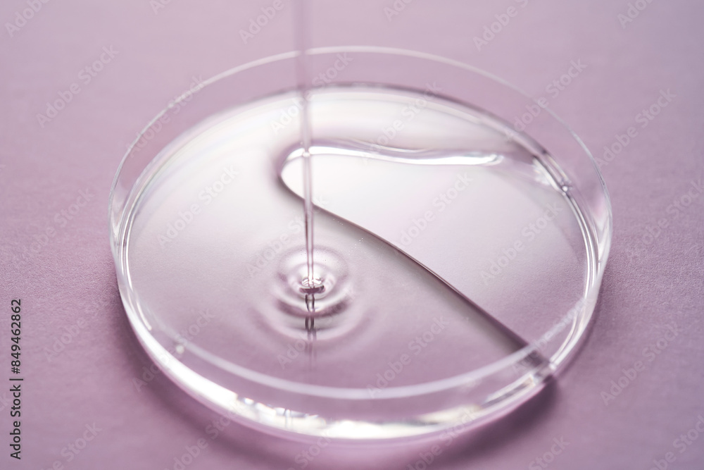 Serum or cosmetic oil flows into a transparent bowl on a purple background. Serum or cosmetic oil flows into a transparent bowl on a purple background.