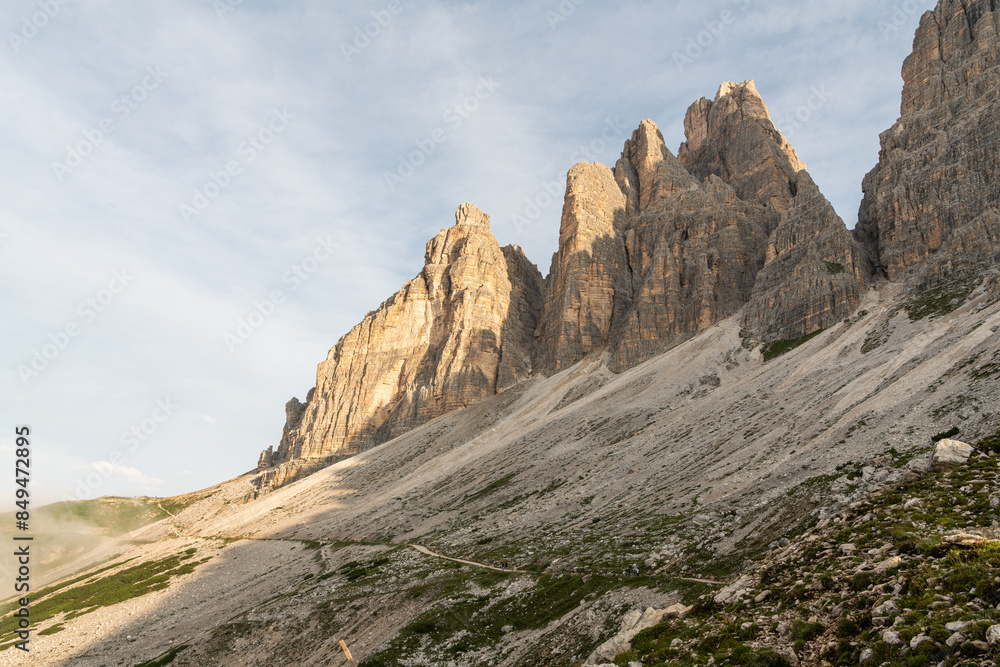 Early morning view of a hiking trail next to impressive rocky mountain peaks towards the Drei zinnen, Tri Crime. The hiking trail is flat, at the side of a steep mountain slope Early morning view of a hiking trail next to impressive rocky mountain peaks towards the Drei zinnen, Tri Crime. The hiking trail is flat, at the side of a steep mountain slope