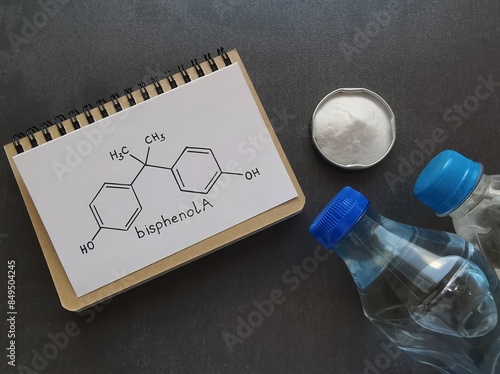 Structural chemical formula of bisphenol A written in notebook with white bisphenol A powder and plastic bottles. Bisphenol A is a chemical compound used in the manufacturing of various plastics.