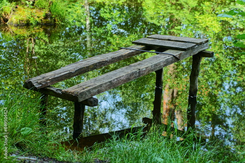 one old homemade wooden primitive bridge broken from the boards for fishing stands in the water of a clean pond near the shore with green grass during the day in summer