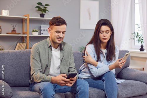 Woman secretly looks suspiciously at screen of man's smartphone, spying on his texting. Couple is sitting on sofa with smartphones in hands. Concept of privacy and relationship dynamics in digital age photo