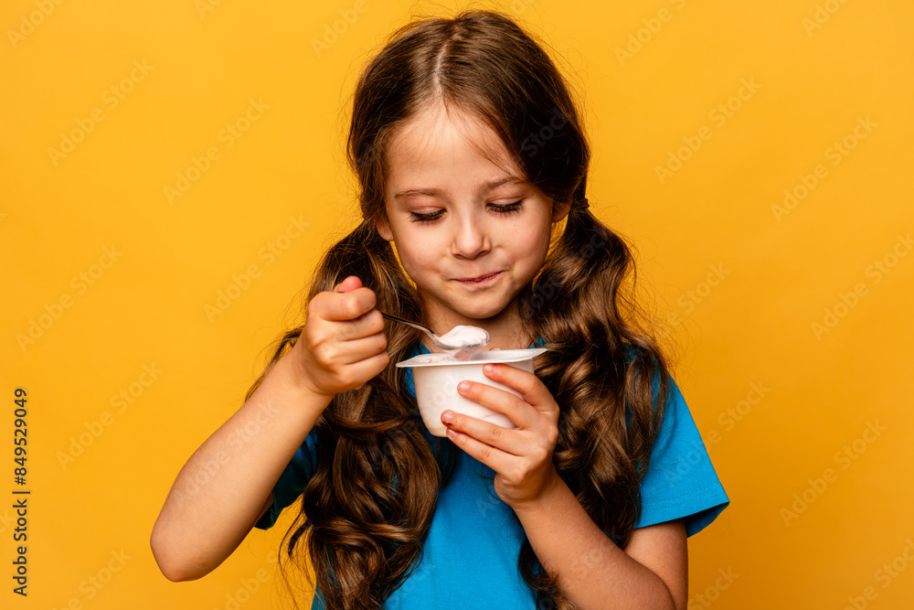 Cute little brunette girl in blue t-shirt eating tasty yogurt on light yellow background. Healthy nutrition concept. Copy space. Cute little brunette girl in blue t-shirt eating tasty yogurt on light yellow background. Healthy nutrition concept. Copy space.