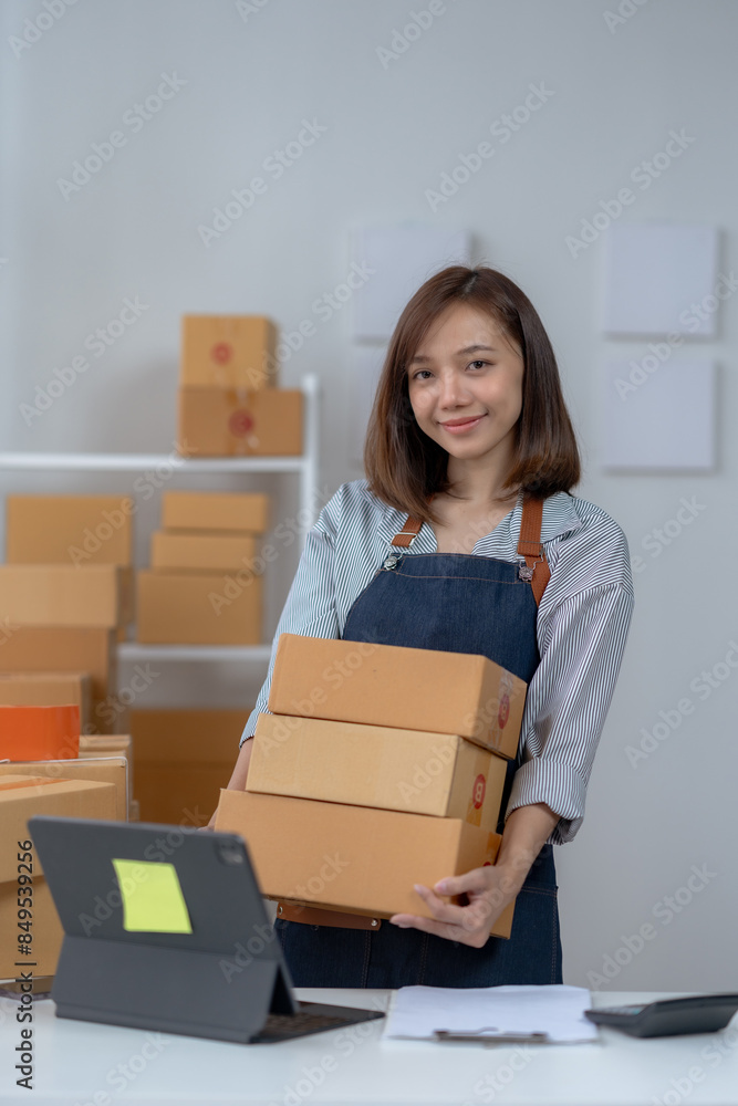 Young woman in apron holding cardboard boxes, working in home office. E-commerce small business concept. Smiling confidently with laptop nearby. Young woman in apron holding cardboard boxes, working in home office. E-commerce small business concept. Smiling confidently with laptop nearby.