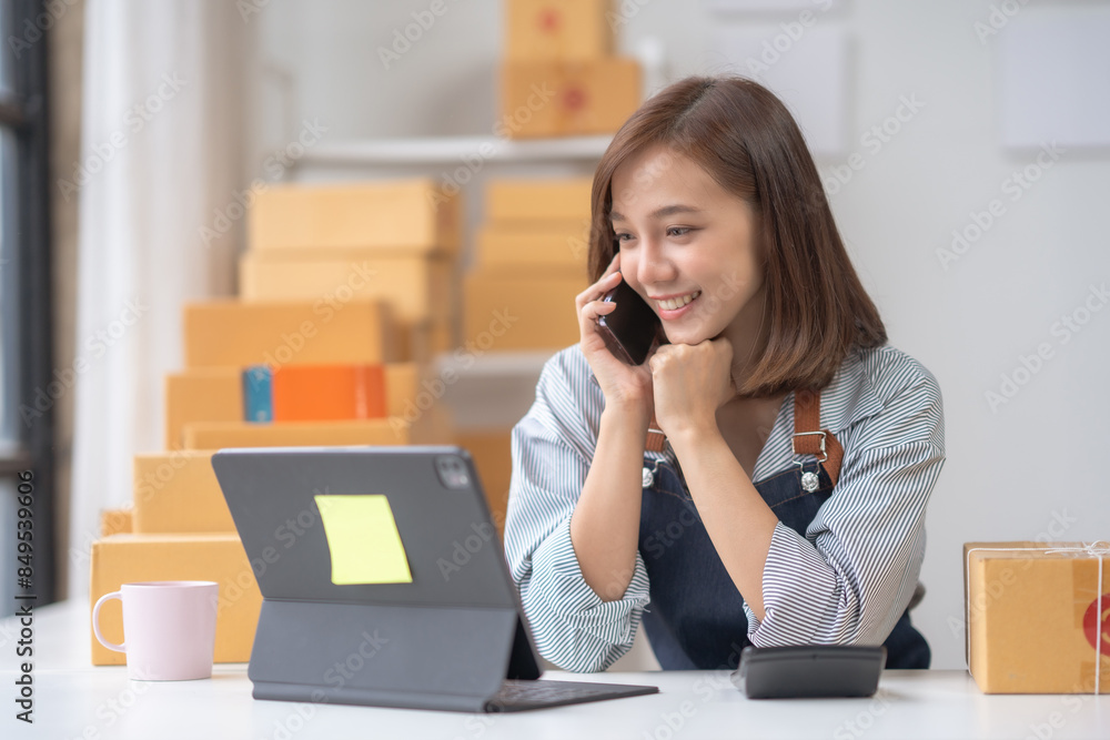 Young woman managing online business, talking on phone, and working on laptop, surrounded by boxes in a modern workspace. Young woman managing online business, talking on phone, and working on laptop, surrounded by boxes in a modern workspace.