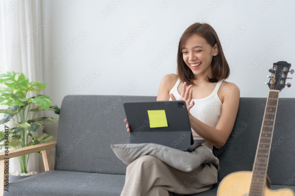 Smiling woman sitting on sofa using tablet for video call, casual indoor setting with a guitar in the background. Smiling woman sitting on sofa using tablet for video call, casual indoor setting with a guitar in the background.