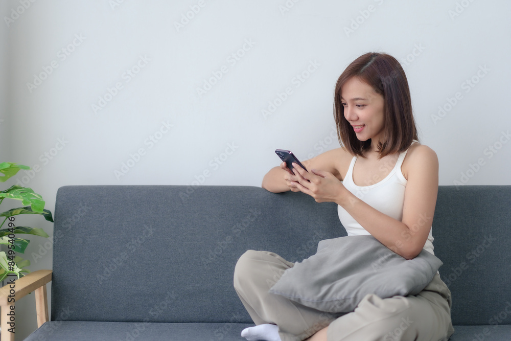 Woman sitting on a grey sofa, smiling while using her smartphone in a relaxed, indoor setting with minimalistic decor. Woman sitting on a grey sofa, smiling while using her smartphone in a relaxed, indoor setting with minimalistic decor.