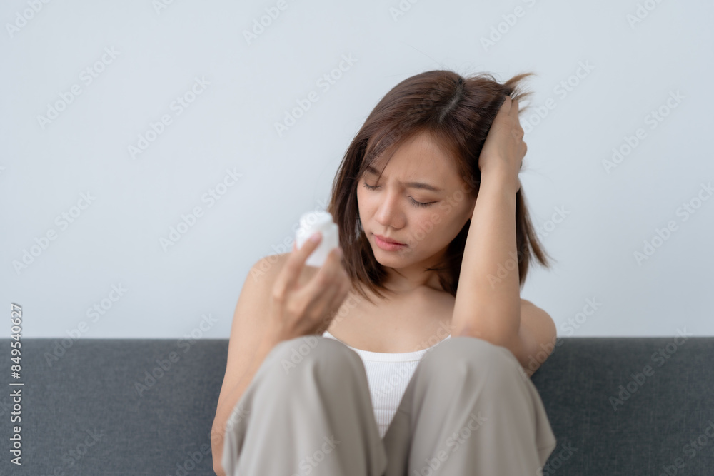 Young woman looking at a pill bottle with a pensive expression, sitting on a sofa and holding her head in concern. Young woman looking at a pill bottle with a pensive expression, sitting on a sofa and holding her head in concern.