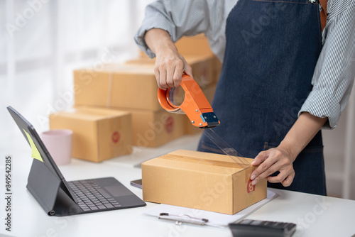 Person sealing a cardboard box with tape beside a laptop, preparation for shipping and delivery, business or e-commerce concept.