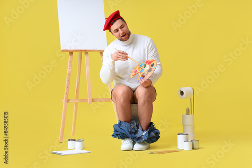 Male artist with paint palette and brush sitting on toilet bowl against yello...