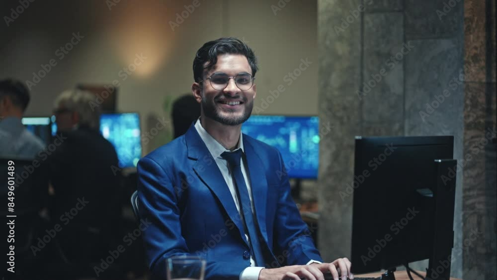 Handsome Caucasian young man working at computer in modern business office. Successful worker cheerfully smiling at camera. People talking with each other and using computers in background. Handsome Caucasian young man working at computer in modern business office. Successful worker cheerfully smiling at camera. People talking with each other and using computers in background.