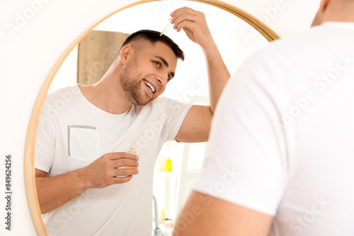 Young man using serum for hair growth near mirror in bathroom