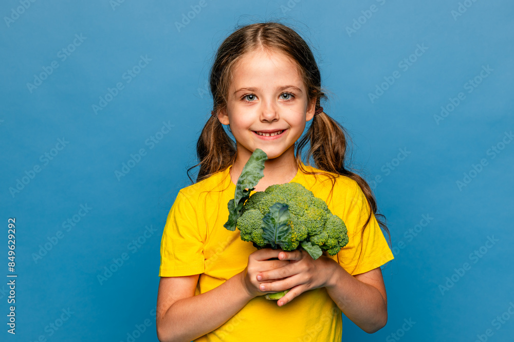 Happy smiling little girl in yellow t-shirt standing with green raw broccoli over blue background. Healthy food for children concept Happy smiling little girl in yellow t-shirt standing with green raw broccoli over blue background. Healthy food for children concept