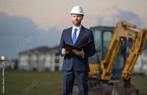 Construction business owner. Man in suit and hardhat halmet at building construction site. Civil engineer worker in front of house near excavator. Architect, supervisor investor, construction manager.