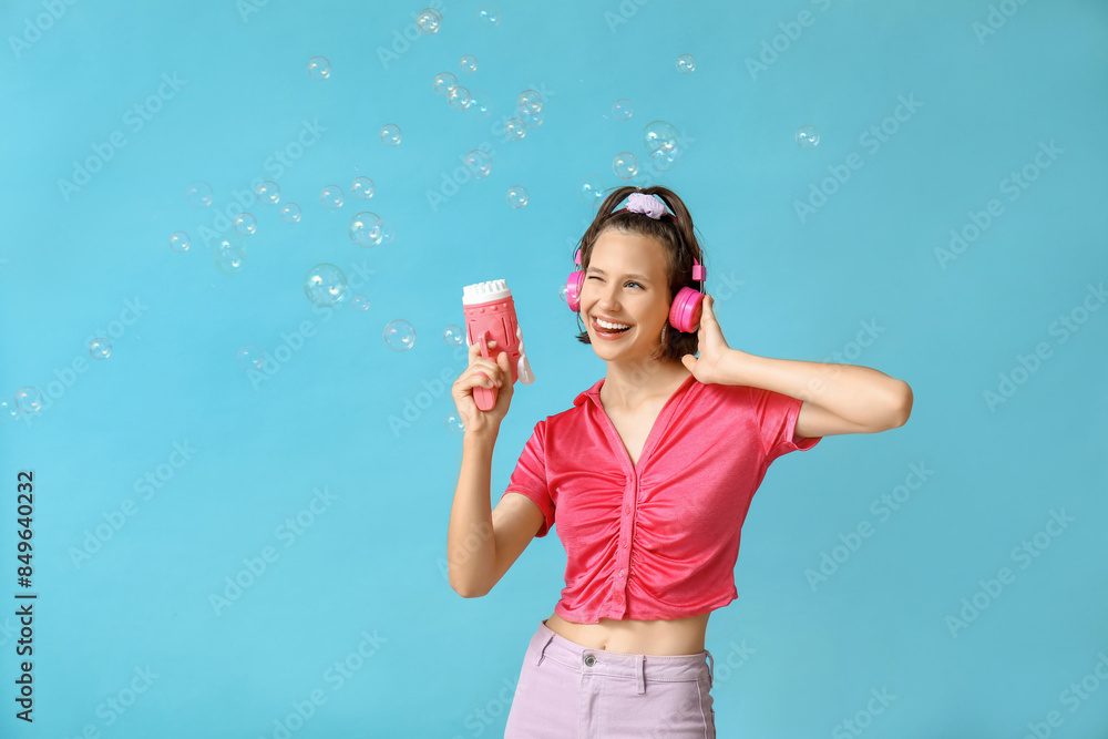 Teenage girl in headphones showing tongue with soap bubble gun on blue background Teenage girl in headphones showing tongue with soap bubble gun on blue background