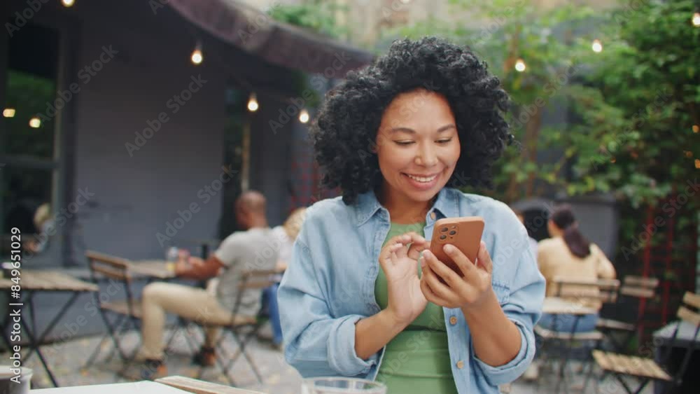 Zooming out from African American woman smiling while scrolling social media on her cell phone. Young female resting with technology gadget in cafe. Having fun while chatting on mobile phone. Zooming out from African American woman smiling while scrolling social media on her cell phone. Young female resting with technology gadget in cafe. Having fun while chatting on mobile phone.