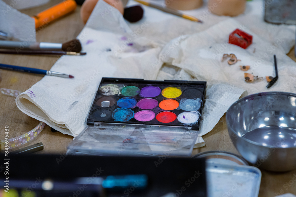 Messy colorful creative table with makeup palette, paints, brushes, pencil sharpener, paintbrushes set and bowl of water, paper tissues on wooden table. Stage make up. Creative chaos. Selective focus Messy colorful creative table with makeup palette, paints, brushes, pencil sharpener, paintbrushes set and bowl of water, paper tissues on wooden table. Stage make up. Creative chaos. Selective focus