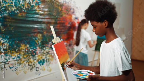 Closeup of african boy hand paint canvas with warm tone color while caucasian girl in casual outfit drawing cool tone colors behind at colorful stained wall. Creative activity concept. Edification.