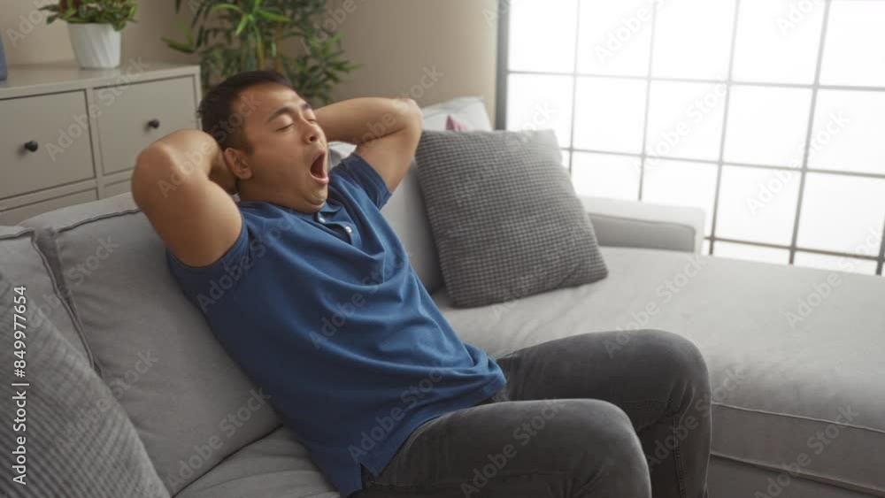 A young chinese man yawning while sitting on a comfortable gray sofa in a bright, modern living room, showcasing a relaxed indoor home environment with contemporary decor. A young chinese man yawning while sitting on a comfortable gray sofa in a bright, modern living room, showcasing a relaxed indoor home environment with contemporary decor.