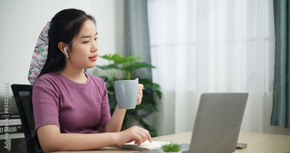 Portrait of Young asian business woman wear wireless headphones drink coffee while using laptop for video conference call presenting project plan to colleagues at home office Portrait of Young asian business woman wear wireless headphones drink coffee while using laptop for video conference call presenting project plan to colleagues at home office