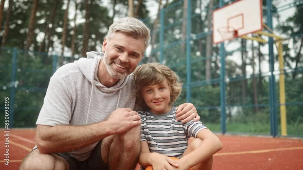 Close up of happy positive family father and little son sitting on basketball court outdoors looking and smiling to camera. Joyful cute kid with ball outside in city park with adult middle-aged dad. Close up of happy positive family father and little son sitting on basketball court outdoors looking and smiling to camera. Joyful cute kid with ball outside in city park with adult middle-aged dad.