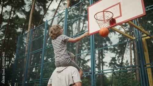Rear of Caucasian family dad and kid boy having fun playing basketball. Happy son sitting on father's shoulders throwing a ball in hoop outdoors on court. Leisure activity. Bottom view. Sport concept