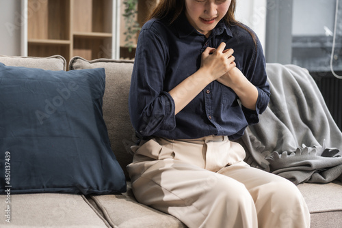 A young female Asian is sitting on a sofa, experiencing a heart attack. She has chest pain, shortness of breath, and radiating pain to her left arm, indicating an emergency. photo