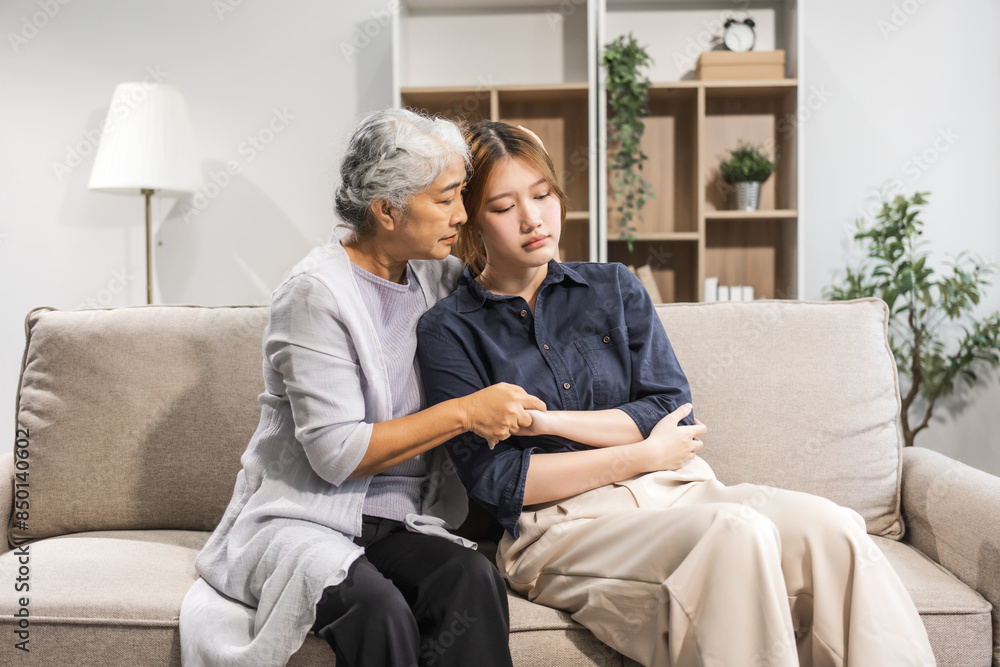 A mature mom and her young daughter, both Asian, sit on a sofa. The daughter appears sick and unwell, causing worry and possibly depression. The mom tries to encourage her and stay by her side.