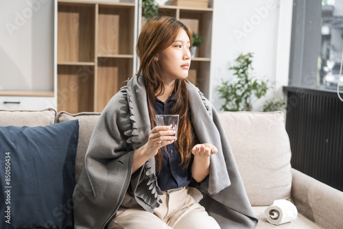 A young female Asian is sitting on a sofa, wrapped in a shawl. She is experiencing cold symptoms like a cough, sneeze, sore throat, and congestion, indicating a viral infection. photo