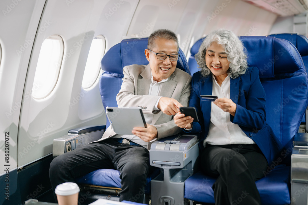 Smiling senior couple of Asian tourists take photos of each other while sitting on a plane. with digital tablet, laptop and smartphone. Travel in style, work Smiling senior couple of Asian tourists take photos of each other while sitting on a plane. with digital tablet, laptop and smartphone. Travel in style, work