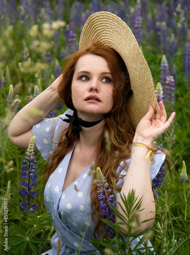 portrait of romantic beautiful readhead girl in blue dress and straw hat  the field of lupines flowers in the summer evening holding lantern