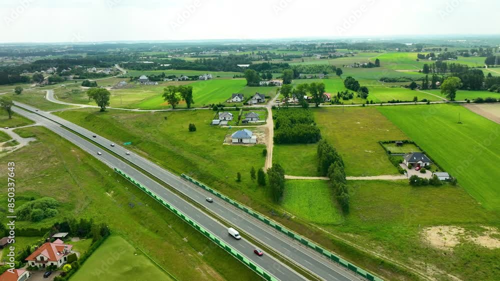 Aerial view of a highway cutting through rural countryside with adjacent residential areas. Cars traveling along the road with lush green fields and scattered homes Aerial view of a highway cutting through rural countryside with adjacent residential areas. Cars traveling along the road with lush green fields and scattered homes