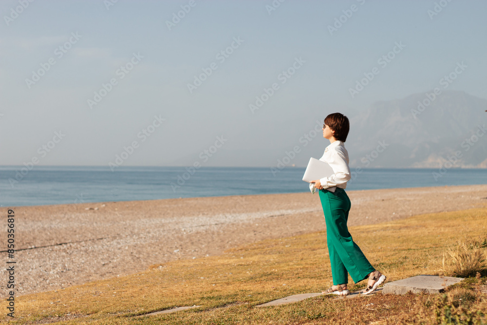 Digital nomad woman traveling the world working with laptop, a girl in a white shirt, green pants, a short haircut goes to the beach with a laptop in her hands Digital nomad woman traveling the world working with laptop, a girl in a white shirt, green pants, a short haircut goes to the beach with a laptop in her hands