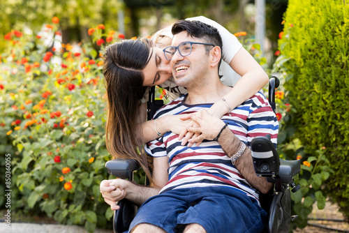 An adult man with a disability sits in an automatic wheelchair with a young woman in a park at sunset.The woman kisses the disabled man on the face.Unconditional support for people with disabilities.