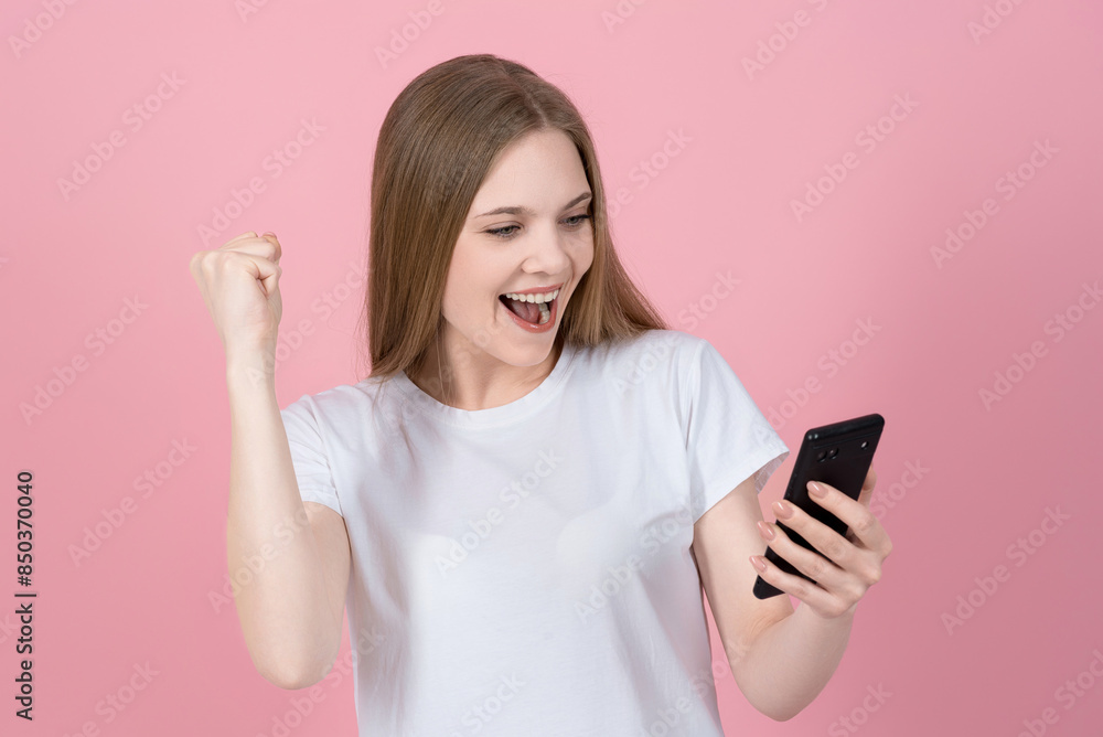 Attractive caucasian young blonde woman in casual white t-shirt shouting happily, using mobile phone and showing winner gesture isolated on pink studio background. Attractive caucasian young blonde woman in casual white t-shirt shouting happily, using mobile phone and showing winner gesture isolated on pink studio background.