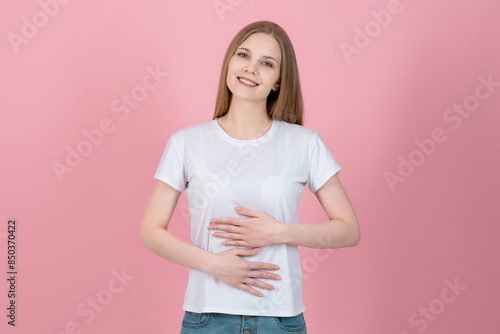 Attractive caucasian young blonde woman in casual white t-shirt looking at camera, smiling and rubbing her belly isolated on pink studio background. Healthy food, delicious, slim.