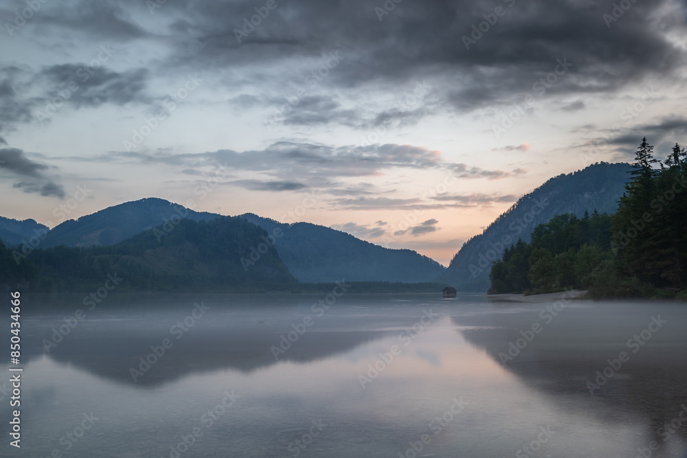 Fototapeta premium A picturesque view of Almsee Lake with Wooden Hut in Austria at sunset. The sky is filled with beautiful, colorful clouds, and the mountains in the distance are reflected in the calm water. 